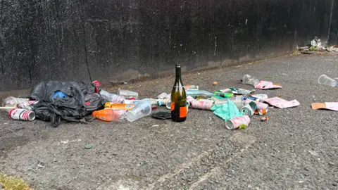 A collection of litter sits on a black concrete pavement. The litter includes cans, bottles, plastic cups and tissue paper.