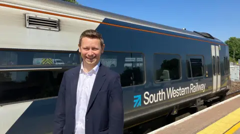 Lawrence Bowman has short light brown hair and is wearing a blue blazer and white shirt. He is standing in front of a South Western Railway liveried train.