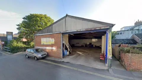 Bubbles Car Valeting. The building is an old garage building, which is empty inside. It has a corrugated iron roof and brick walls. A yellow banner advertises it as a car wash.