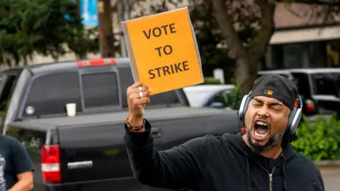 Getty Images A man wearing a baseball cap backwards and headphones on shouts and holds up a placard saying 'vote to strike'.