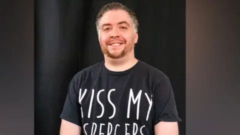 Mark Nicholas A man smiles at the camera with a black background and black top. He has a short beard and hair. 