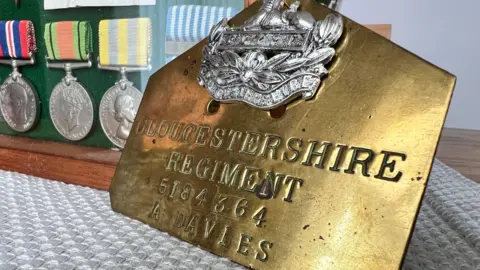 A gold metal plate stamped with the words' Gloucestershire Regiment, A. Davies'. Next to it are three silver medals with coloured stiped straps displayed in a glass case. 