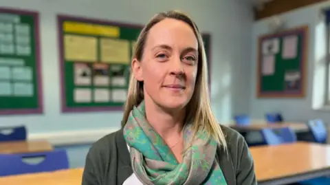 A close-up of Amy in a classroom, smiling to camera, wearing a muted turquoise and lilac scarf and a dark green cardigan