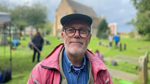 Stephen Driscoll wears a cap, glasses and a red jacket as he stands in the churchyard 