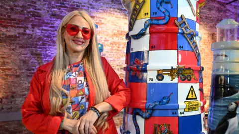 St Luke's Hospice Plymouth/Paul Slater Images Mandi has blonde hair and is wearing a red jacket and sunglasses, standing next to a lighthouse sculpture which looks like a snakes and ladders board