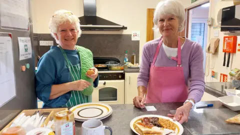 Devizes Opendoors Two older women with grey hair stand in a kitchen setting behind a plate of toast, beans, bacon and mushrooms.
