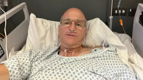 David Stoyle A selfie taken by David whilst he is laying in a hospital bed wearing a hospital gown.