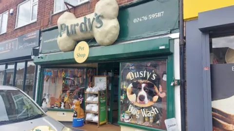 The frontage of a pet food shop. A large sign in the shape of a bone that says Purdy's Pet Shop sits above the door. The window frames are painted green, on the right window is a picture of a dog in a lying/begging position that says Dusty's Dog Wash.