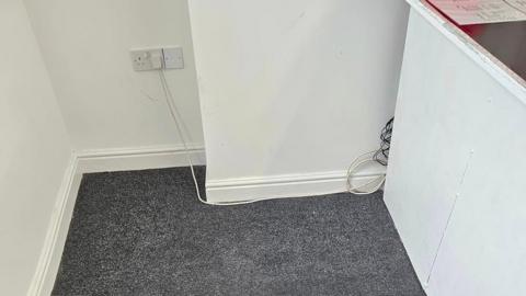 The corner of an empty room. The carpet is grey and the walls are white