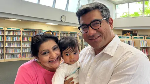 Aasim Hussain on the right holding his toddler son Armaan in the middle of the shot, and his son’s grandmother Fouzia is on the left. They are in Brompton Library in Kensington