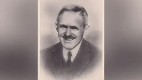 Susan Basnett Thomas Roby with a moustache, greying hair in a suit & tie and white shirt.