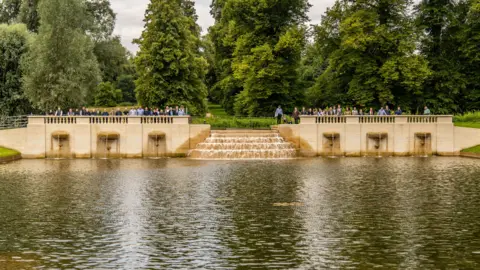 Boughton House Star Pond, showing a stone structure, with water at the front, as small waterfall and a large amount of people standing on the wall. There are large trees behind them all. 
