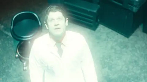 Out There A brunette man in a white shirt, black tie and white lab coat looking up at a light which is shining on him.