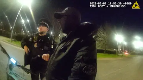 Essex Police A person wearing black with a black balaclava covering their face and a black baseball cap standing side on in the picture. A police officer is next to him and holding the individual's wrist. The handle bars of an electric bike can be seen next to them. it is at night. The image is from a body worn camera.