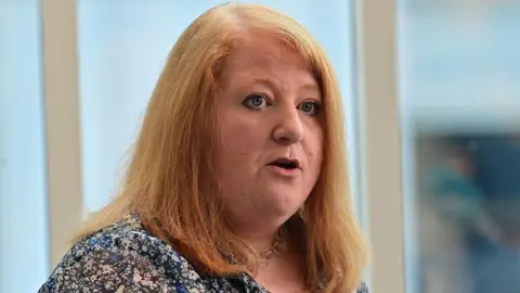 Naomi Long has long red hair and is wearing a flowery blouse
