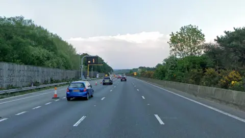 A stretch of motorway. It is lined with trees and the sky is overcast. 