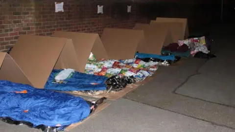 LIGHT PROJECT PETERBOROUGH A line of sleeping bags with cardboard on top next to a bare brick wall on concrete ground. There is cardboard underneath the sleeping bags. One of the sleeping bags is made of a patchwork of crisp packets. 