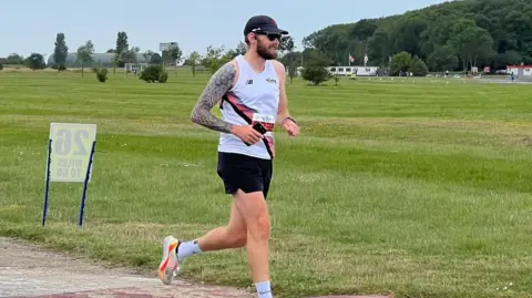 Brain Tumour Research Alex with short dark hair and beard, dressed in running kit, running past a large area of grass with trees and a mountain in the background. He has just passed a 26 mile marker.