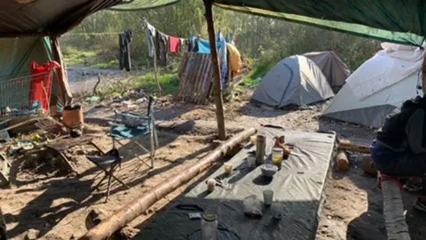 There is an ottoman table with cups in the foreground, a tent neatly pitched behind, and laundry hanging on a rope