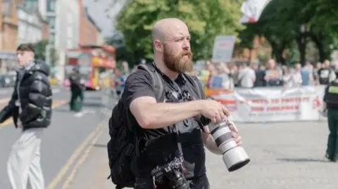 Brendan Harkin Brendan has a bald head with a beard. He has a black t-shirt and is holding a camera.