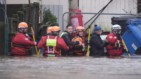 PA Media Seven men, dressed in red search and rescue clothing, hi-viz vests and helmets, are wading in waist-high water