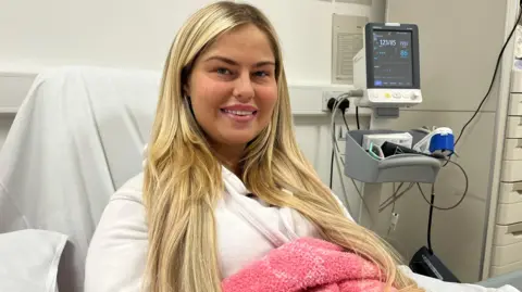 Kenzi Quinn smiles at the camera while receiving dialysis treatment at Sunderland Royal Hospital's Renal Unit. Kenzi has blonde hair and is wearing a white hooded jumper while wrapped in a pink and white blanket.