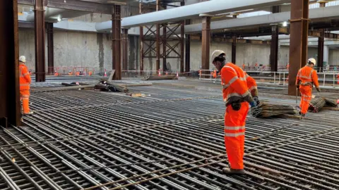 Three workers stand on a grid of steel. They are inspecting the work.