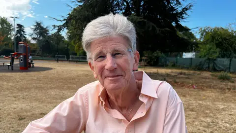 Steve Allen, a white man in his 70s with grey hair and unframed glasses smiles at the camera. He's in a button up pink shirt with the top button undone. He is sat in a park with browned grass and a tree behind him.