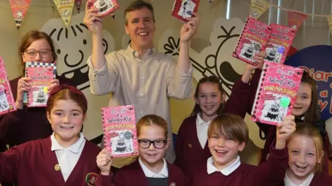 Owen Humpreys/PA Kinney is at the back smiling and holding up copies of his books. He is with seven smiling children in maroon jumpers also holding up his books. In the background are cartoons of the wimpy kid from his stories. 