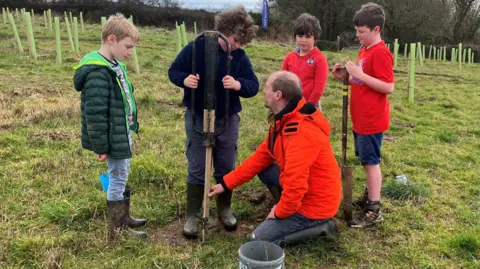 Four young boys stand around while Hastings with dark curly hair holds the stamper to push in a piece of baton next to the sapling that will protect the plant 