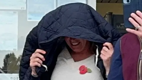 A close-up photo of Rebecca Pinckard walking out of court with her black coat over her head as she tries to cover her face. She appears to be smiling under the coat. She is wearing a cream jumper with a poppy on it.
