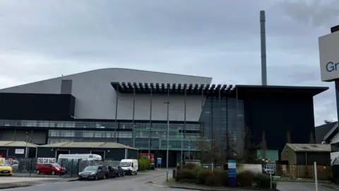 BBC News A large building with a chimney to the right hand side. It is covered in grey panels and has large windows on the first four floors. It is set against a grey sky. There is a car park in the foreground