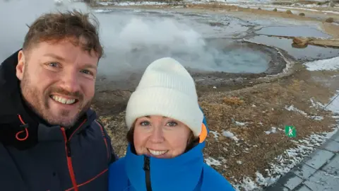 A man and a woman wearing warm winter clothing stand in front of steam rising from the ground in Iceland