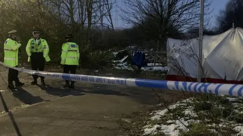 Three police officers, wearing yellow hi-vis jackets, stand behind police tape which cordons off the area in which a baby's remains were found. 