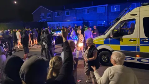 A crowd of people gathered outside an address in Thetford. There is also a marked police van to the right of the picture.