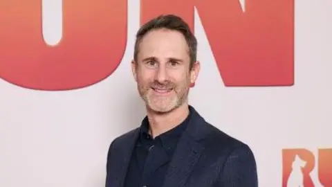 A grey-haired man wearing a dark blue suit smiles at the camera.