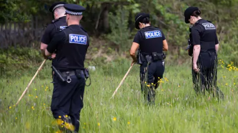 Four police officers, two men and two women, in black uniforms stand with their backs to the camera in a patch of long grass carrying wooden sticks used to search for weapons or drugs. 
