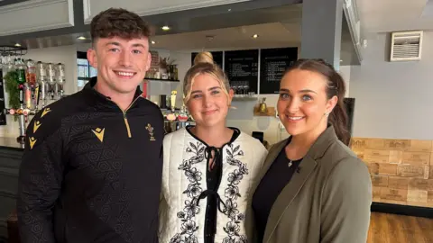 An image of me, Finley and Bronnie stood in front of a bar at our local pub. We are all smiling looking at the camera. Finley, on the left, is wearing a black quarter zip sports top. He has short brown hair. I am in the middle, I am wearing a cream and black padded patterned jacket. My hair is in a top bun. Bronnie, on the right, is wearing a green blazer and black top. Her brown hair is in a high ponytail.