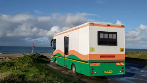A picture of the coach in which the library will be housed. It's parked on the side of the road looking out to sea. 