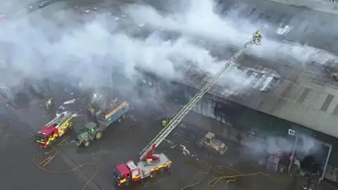 A number of fire appliances, including one with an extended ladder battle a fire at a large recycling plant. Thick grey smoke is flowing from the building which has a grey slanted roof.