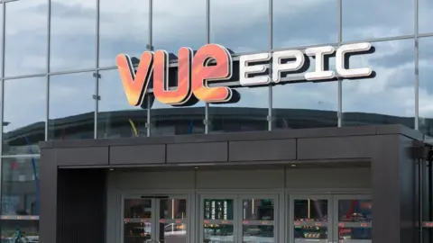 A close-up of a sign fixed to a glass building, which reads VUE EPIC. Below the sign are the modern glass doors into the cinema building.