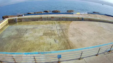 An empty concrete pool with a 'NO DIVING' warning painted near the bottom edge. A metal railing surrounds the pool, and a ladder is visible on the far side. In the background, the ocean stretches out with several rocks protruding from the water.
