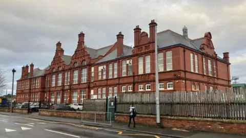 Large red brick building behind gates. A road and crossing can be seen in the foreground of the photo. 
