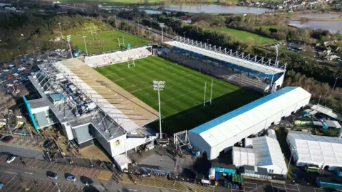 Aerial shot of Sandy Park in Exeter on a sunny day. The pitch is surrounded by four stands with a large car park to the left of the stadium and a large river to the right.