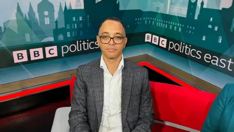 Nic Rigby/BBC Tim Roberts is sitting on a red chair in a TV studio. In the background it says "BBC Politics East". He is wearing spectacles and a pale shirt and grey jacket.
