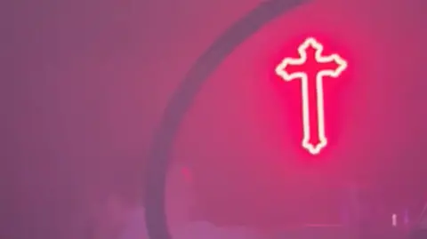 A light in the shape of a pink neon cross hanging in a night club 