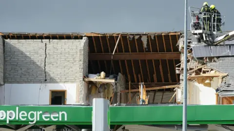 PA Media Shows an Applegreen service station with a hole in the rood and members of the emergency services looking down on it to the right from an extending ladder.