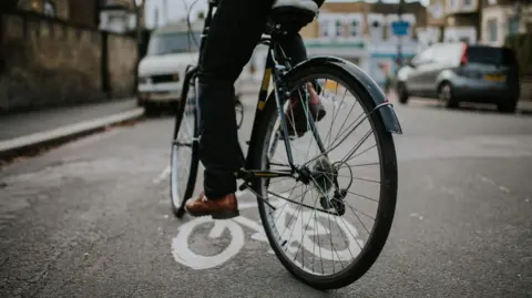 Getty Images A person riding a bicycle, only their legs are visible in the shot. They are on a street with some cars visible