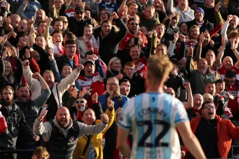 Tom Banks/MFC A large crowd of people cheer as a footballer in a blue and white striped shirt faces them