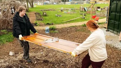 EARNT Two people carrying a plank of wood outside.